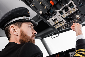 bearded pilot in cap reaching overhead panel with set of switches in airplane simulator