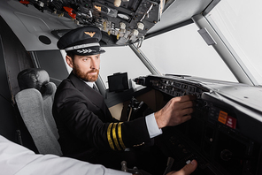 pilot in cap and uniform reaching control panel near co-pilot in airplane simulator