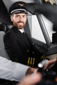 bearded pilot in uniform and cap smiling near blurred co-pilot