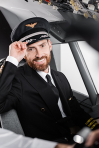 bearded pilot in uniform adjusting cap and smiling near blurred co-pilot