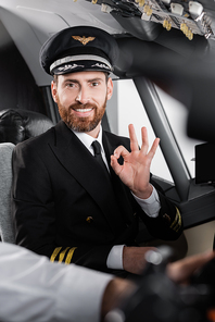 bearded pilot in uniform showing okay and smiling near blurred co-pilot