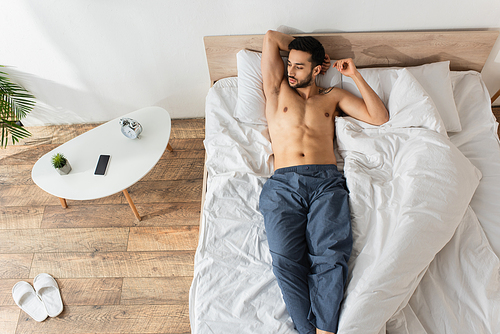Top view of shirtless man lying on bed near smartphone with blank screen and alarm clock on bedside table