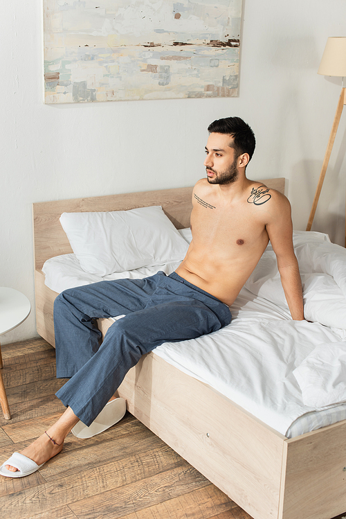 Shirtless man in pants and slippers sitting on bed