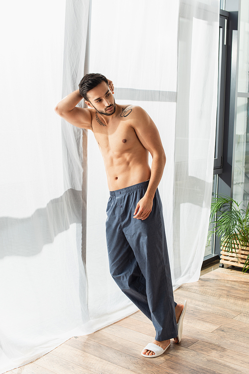 Shirtless man in slippers and pants standing near window