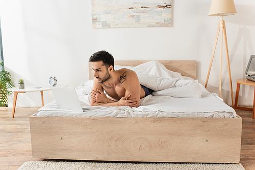 Shirtless man looking at laptop on bed in morning