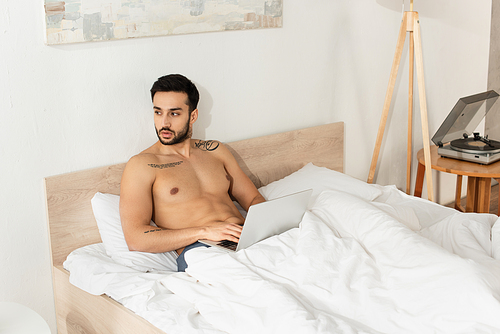 Shirtless freelancer using laptop on bed