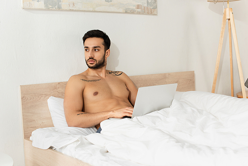 Muscular teleworker using laptop while sitting on bed in morning