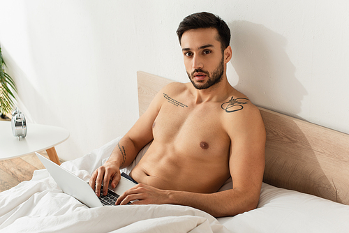 Shirtless freelancer using laptop on bed near alarm clock on bedside table