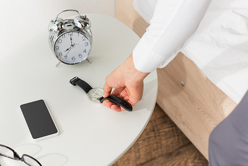 Cropped view of businessman taking wristwatch from bedside table near smartphone and eyeglasses