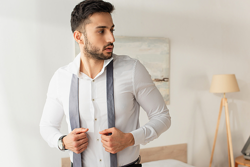 Bearded businessman in shirt holding tie at home
