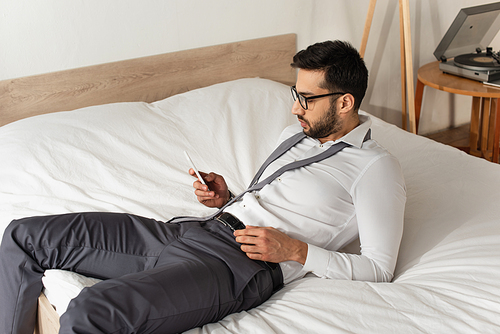 Side view of bearded businessman using smartphone on bed