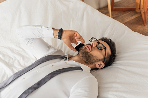 Businessman in eyeglasses and shirt talking on smartphone while lying on bed