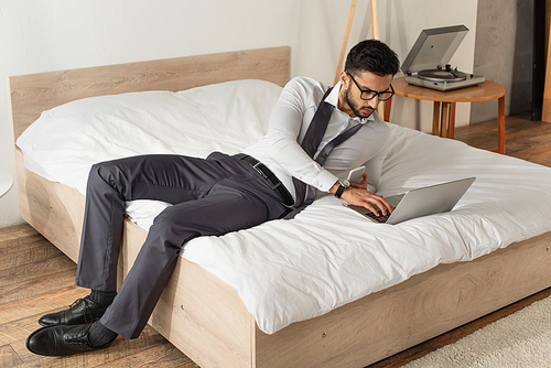 Businessman with smartphone using laptop on bed at home