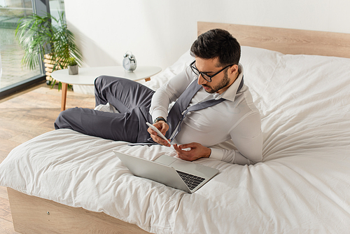 Businessman using smartphone near laptop on bed at home