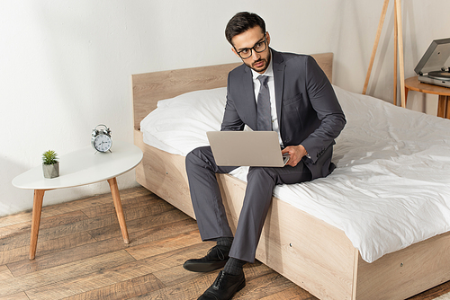 Businessman in eyeglasses holding laptop on bed near alarm clock on bedside table