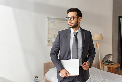 Young businessman with laptop standing in bedroom at home
