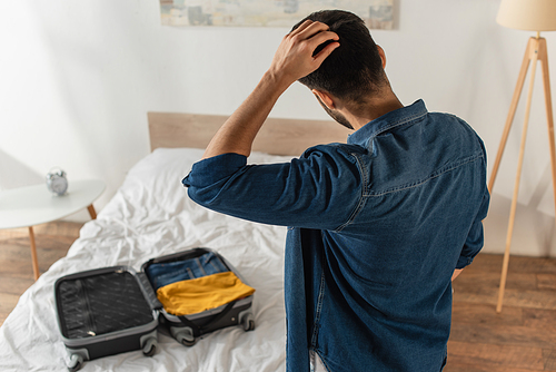 Back view of man holding hand near head standing near suitcase on bed