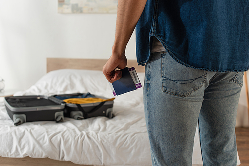 Cropped view of man with passport and boarding pass standing near blurred suitcase in bedroom