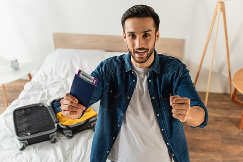 Excited man holding passport and showing yes gesture near blurred suitcase on bed