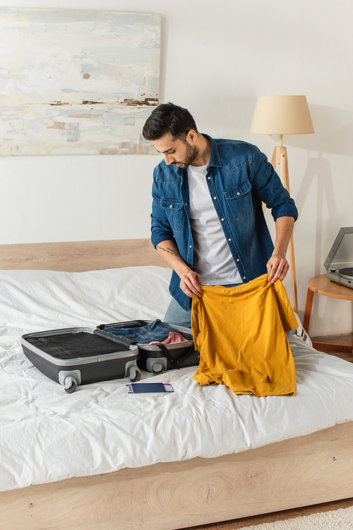 Young man holding t-shirt and looking at suitcase near passport on bed