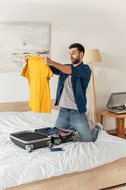 Pensive man looking at t-shirt near suitcase and passport in bedroom