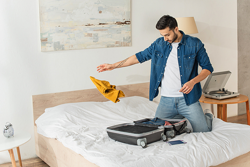 Man throwing clothes near suitcase and passport on bed