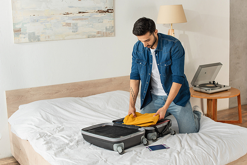 Bearded man putting clothes in suitcase near passport on bed