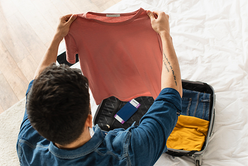 Overhead view of man holding t-shirt near suitcase with passport on bed