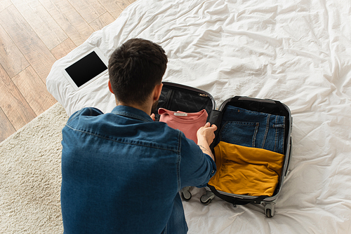 Overhead view of man putting clothes in suitcase near digital tablet on bed