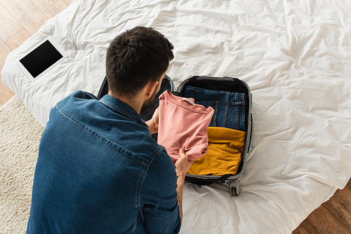 Overhead view of man putting clothes in baggage near digital tablet with blank screen on bed