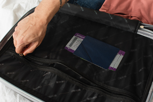 Cropped view of man fastening zipper of suitcase with passport on bed