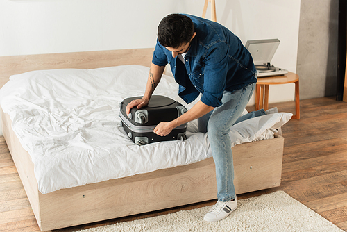 Young man zipping suitcase on bed at home