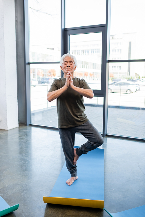 Elderly asian man standing in tree pose on yoga mat in sports center