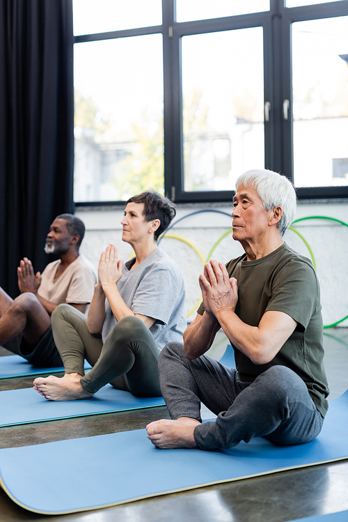 Elderly asian man sitting in yoga pose near multiethnic friends in sports center