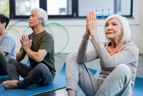 Grey haired woman practicing yoga near blurred asian friend in sports center