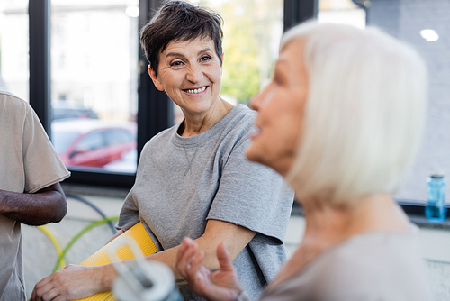 Smiling woman with fitness mat looking at blurred grey haired friend in gym