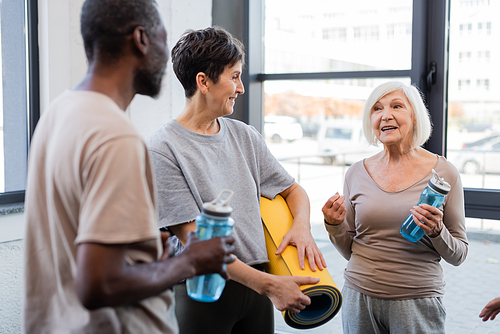 Senior woman holding sports bottle and talking to interracial friends in gym