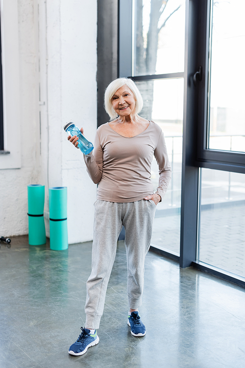 Positive elderly sportswoman holding sports bottle in gym