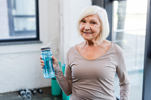 Grey haired sportswoman with sports bottle smiling at camera in gym