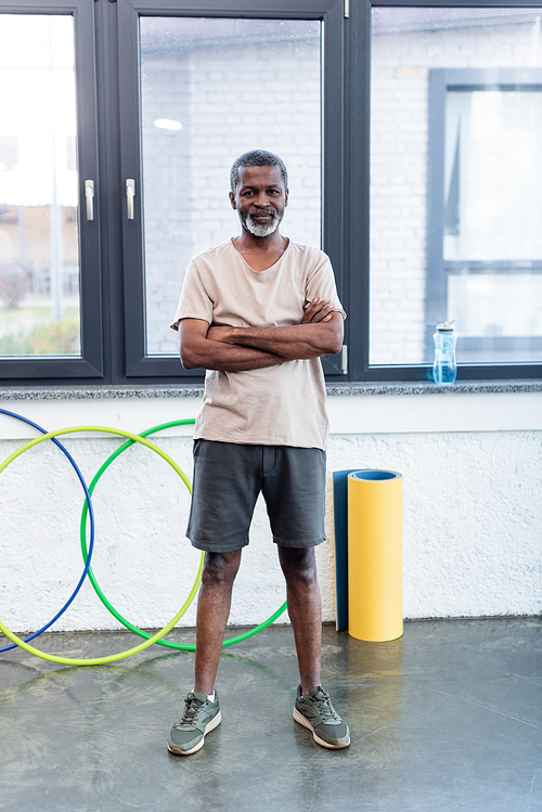 Senior african american sportsman standing with crossed arms near fitness mat and hula hoop in gym