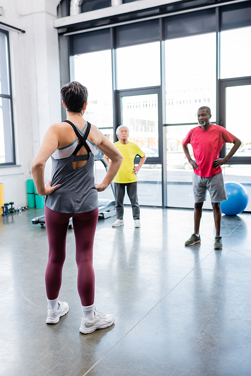 Senior trainer holding hands on hips near blurred multiethnic people in gym