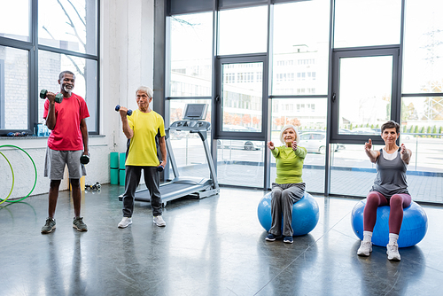 Multiethnic sportsmen training with dumbbells near sportswomen on fitness balls in gym