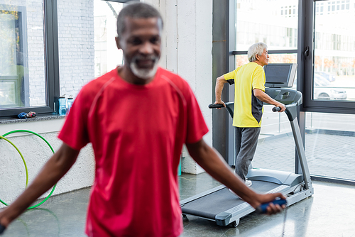 Senior asian man training on treadmill near blurred african american man with jump rope in gym