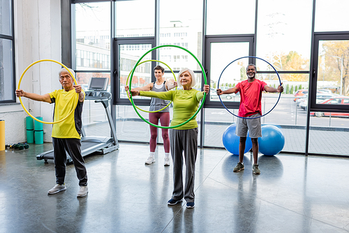Multiethnic senior people training with hula hoops near treadmill and fitness balls in gym