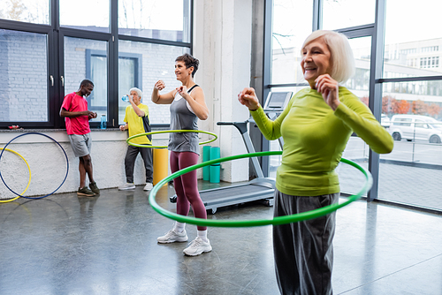 Elderly sportswoman training with hula hoop near friend and interracial sportsmen in gym
