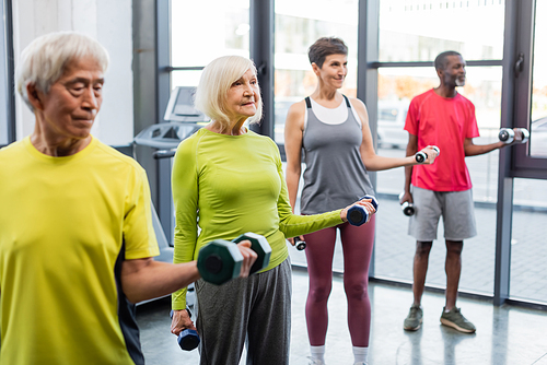 Smiling grey haired woman training with dumbbells near multicultural friends in gym