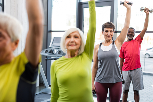 Elderly woman working out with dumbbells near multiethnic friends in gym