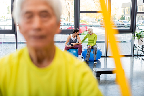 Smiling elderly sportswomen talking on fitness balls near blurred man training in gym