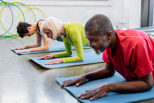 Senior woman exercising on fitness mat near interracial people doing plank in gym