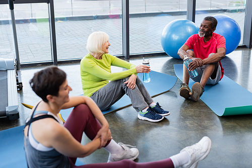Cheerful senior interracial people sitting on fitness mats in sports center